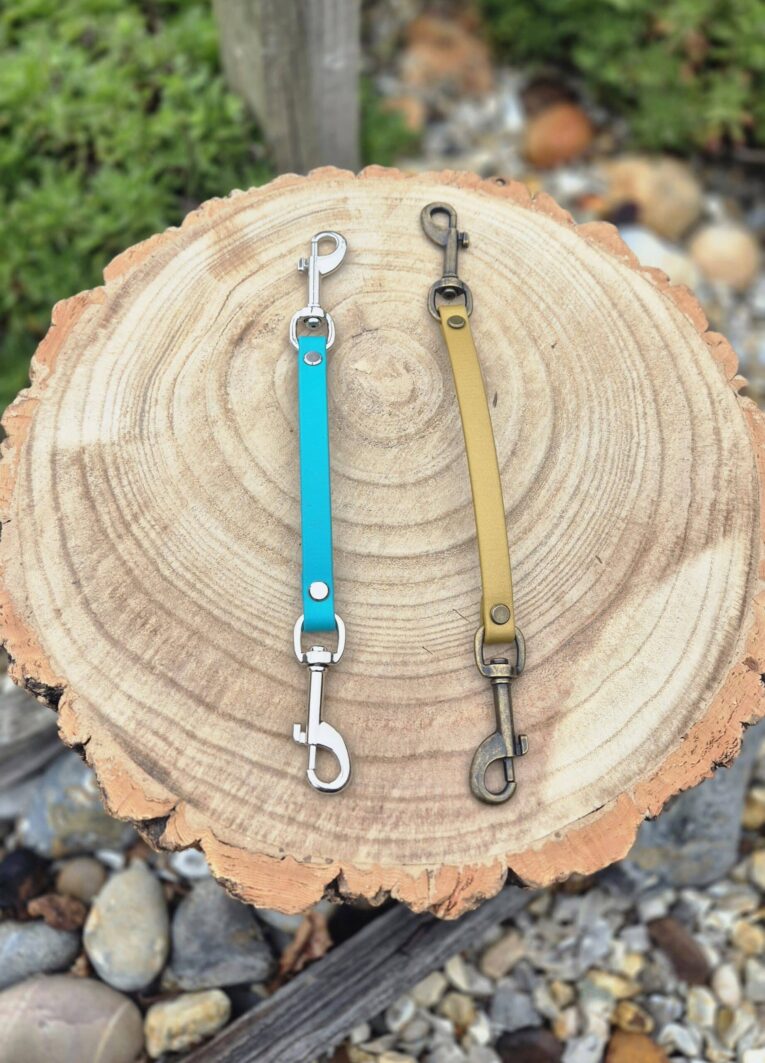 Two BioThane safety clips in turquoise and tan displayed on a wooden log cross-section.