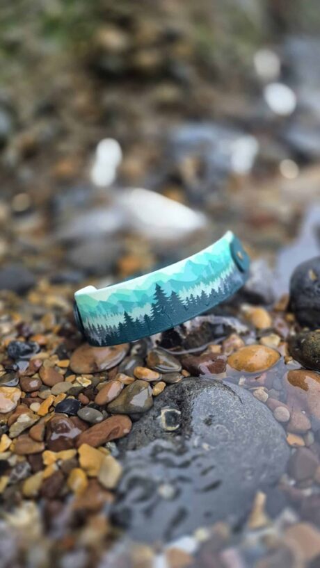Forest print tapered hound collar in turquoise and teal displayed on coastal pebbles and rocks.
